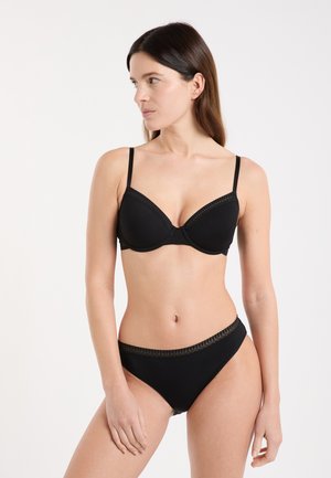 Donna che indossa un reggiseno nero e mutandine abbinate con rifiniture in pizzo, in piedi davanti a uno sfondo bianco uniforme, che guarda di lato.