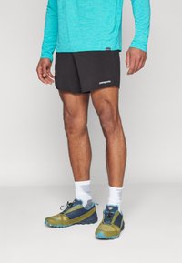 Black athletic shorts with a white "Patagonia" logo, paired with a long-sleeve teal shirt and green and blue trail running shoes.
