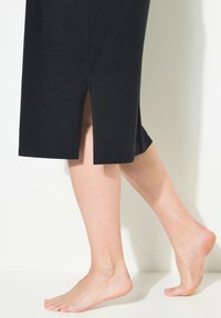 Černé šaty po kolena s bočními rozparky, hladkou texturou, krátkými rukávy a volným střihem, předváděné na barevé noze.