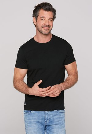 Camiseta negra de manga corta hecha de algodón, con un escote redondo. Combinada con jeans de color azul claro, exhibida sobre un fondo liso.
