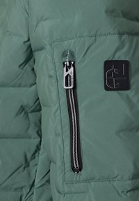 Grüner Steppjackenabschnitt mit vertikaler schwarzer Reißverschlusstasche und schwarzem rechteckigem Logo-Patch mit abstraktem Design.