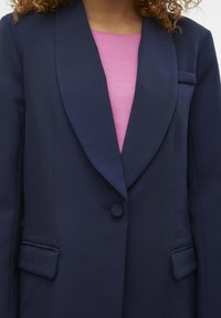 Sacou bleumarin cu rever ascuțit, închidere cu un singur nasture și două buzunare frontale. Purtați peste o cămașă roz deschis. Textură de stofă netedă.