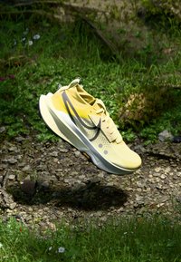 Chaussure de sport jaune clair avec un dessus en maille respirante, accents gris foncé, semelle texturée et détails perforés pour la ventilation.