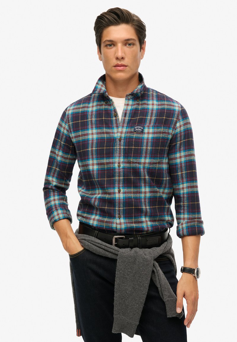 Camicia di flanella con un motivo a quadri blu e verde, maniche lunghe, colletto button-down, con logo sulla tasca del petto. Jeans in denim scuro.