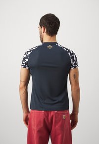 Männer Sportshirt in Dunkelblau mit kurzen Ärmeln. Mit einem geometrischen weißen Muster auf den Schultern und einem kleinen Logo auf dem oberen Rücken.