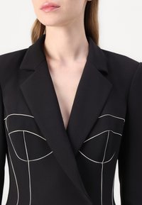 Čierny blazer s bielym kontrastným prešitím, ktorý má hlboký V-výstrih, špicaté klopy a tvarovaný dizajn korzetu. Hladká textúra látky.