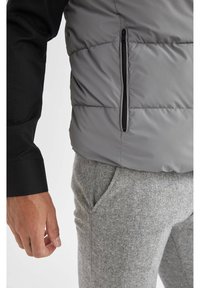 Homme portant une chemise noire à manches longues, un gilet rembourré gris avec une poche zippée verticale et un pantalon de survêtement gris avec une poche latérale.
