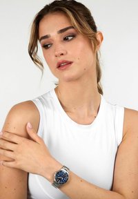 Orologio in acciaio inox argentato con quadrante blu, caratterizzato da indici delle ore argentati, una finestra per la data e un bracciale in metallo. Modello che indossa una maglia bianca.