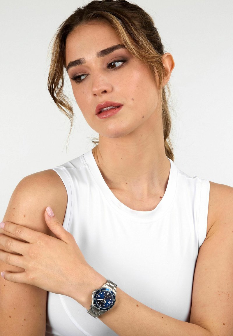 Orologio in acciaio inox argentato con quadrante blu, caratterizzato da indici delle ore argentati, una finestra per la data e un bracciale in metallo. Modello che indossa una maglia bianca.