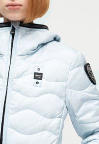 Άτομο που φοράει ανοιχτό μπλε καπιτονέ μπουφάν με κουκούλα, φερμουάρ μπροστά, μαύρο λογότυπο "Blauer U.S.A." στο στήθος και εμβληματικό λογότυπο στο μανίκι.