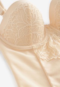 Béžová čipkovaná podprsenka s kvetinovými vzormi, vystuženými košíčkami a nastaviteľným ramienkom na bielom pozadí.
