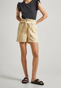 Beżowe szorty z wysokim stanem, paskiem w talii, bocznymi kieszeniami i luźnym krojem. W połączeniu z czarną bluzką z koronkowymi, falbaniastymi rękawami.