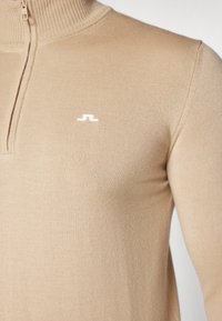 Beige stickad tröja med hög krage och halvlång dragkedja. Har en liten vit logotyp på vänster bröst. Slät textur.