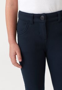Pantaloni elasticizzati blu navy con una texture liscia, dotati di chiusura frontale con bottone, passanti per cintura e tasche laterali. Vista ravvicinata dell'elastico in vita.