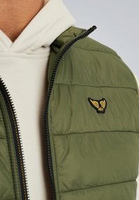 Vadderad grön väst med dragkedja, grå hoodie under, som har en broderad svart och gul logotyp. Texturerad yta med horisontell sömnad.