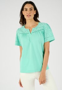 Top a maniche corte in cotone verde menta con motivi a occhio sulle spalle, caratterizzato da una scollatura a V e una vestibilità rilassata.