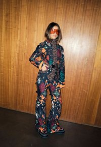 Nyomtatott overál, amely sötét tónusú virágos és absztrakt minták keverékét mutatja, narancssárga részletekkel és masszív fekete lábbelivel kiegészítve.