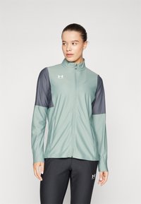 Svijetlozeleni zip-up jakna sa sivim rukavima, izrađena od mekanog materijala. Ima visoki ovratnik i logotip na prsima i rukavu.