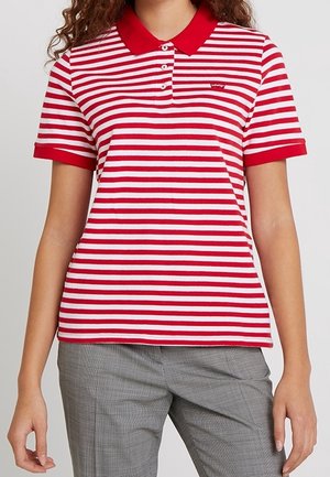 Femme portant un polo à manches courtes rayé rouge et blanc avec un col rouge et un pantalon gris à carreaux.