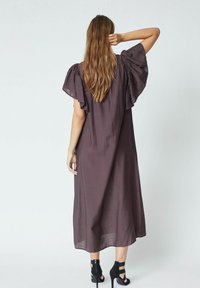 Rochie maxi maro, vaporoasă, cu mâneci supradimensionate și volănașe, dintr-un material ușor și cu o siluetă lejeră. Tocuri vizibile la baza rochiei.