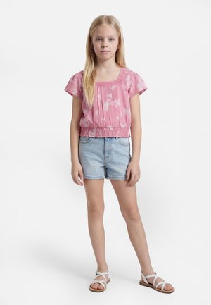 Κοκκινομάλλα κοπέλα στέκεται με το πρόσωπο μπροστά φοράει ένα ροζ μπλουζάκι με κοντά μανίκια, ανοιχτόχρωμα τζιν σορτς και άσπρα σανδάλια σε λευκό φόντο.