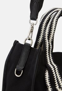 Černá textilní taška s koženým uchycením a černobílým tkaným ramenním popruhem, se stříbrnou sponou s vyrytým nápisem "Karl Lagerfeld."