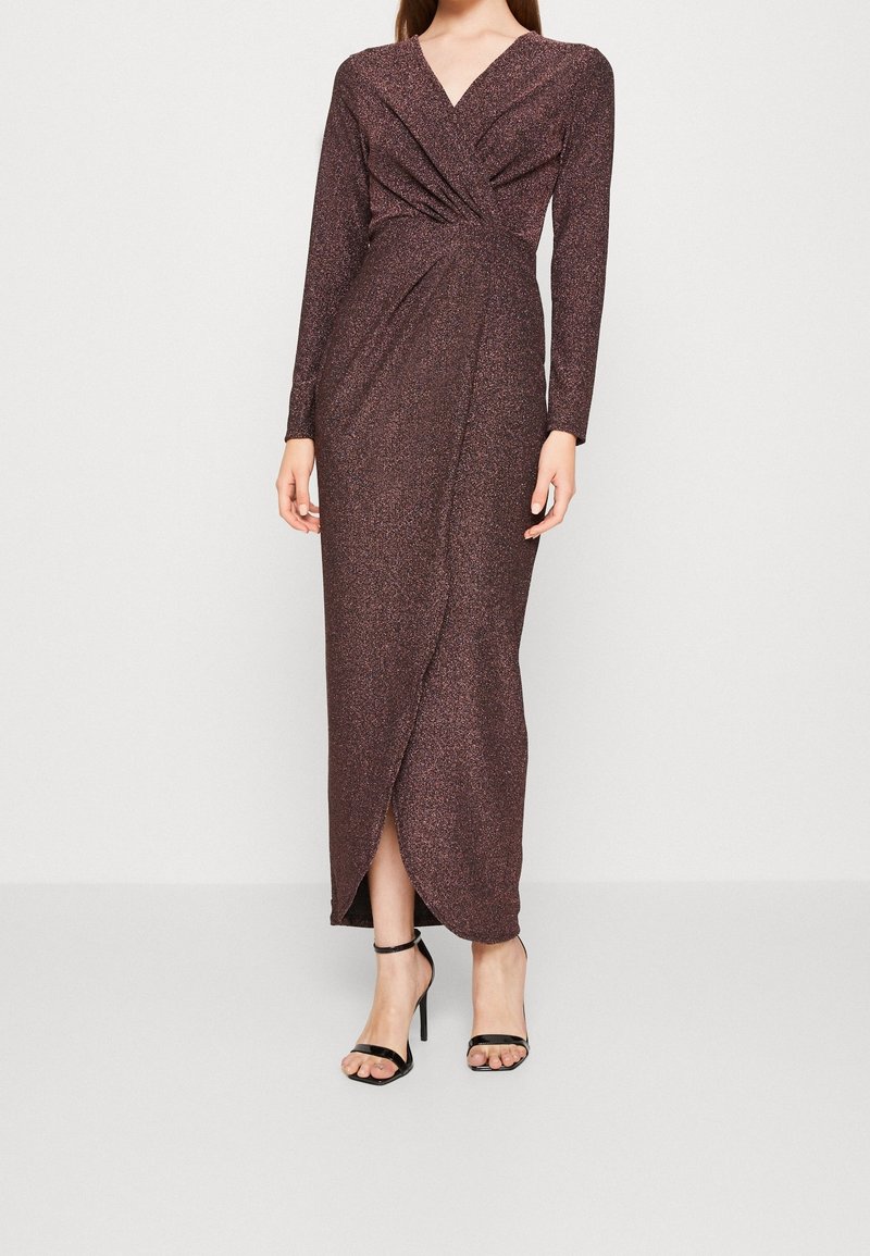 Femme portant une robe portefeuille marron scintillante à manches longues avec un col en V et des sandales noires à talons hauts, debout sur un fond uni.