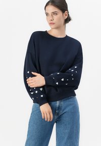 Marineblå genser med oversized passform, med sølvfargede hjerte mønstre på ermene. Rund hals og ribbede mansjetter. Kombinert med lyseblå jeans.