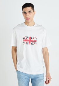 Camiseta de algodón blanca con un estampado desgastado de la Union Jack en el pecho. Manga corta y ajuste relajado. Jeans de mezclilla claros.