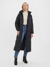 Manteau d'hiver noir long avec capuche, présentant un design matelassé, des zippers latéraux et une coupe mi-longue. Porté par-dessus un pull beige et un jean bleu.