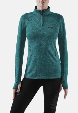 Femme portant un haut de sport à manches longues bleu sarcelle avec fermeture éclair et trous pour les pouces, et des leggings noirs avec un motif sur la cuisse.