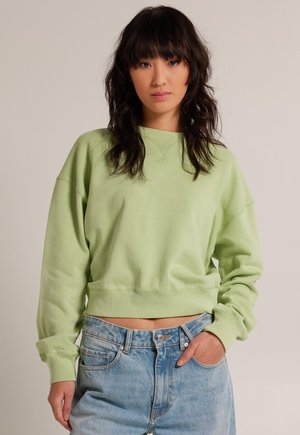 Sudadera verde claro corta con escote redondo, hombros caídos, puños acanalados y textura suave, combinada con pantalones cortos de mezclilla de cintura alta.
