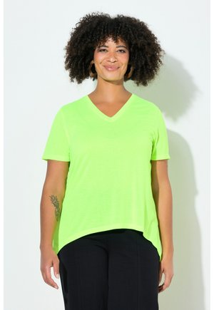 Frau mit lockigem Haar lächelnd, trägt ein hellgrünes T-Shirt mit V-Ausschnitt und schwarze Hosen, steht vor einem weißen Hintergrund.