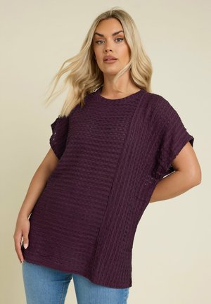 Paarse textuurtop met korte mouwen, een ronde hals en een mix van horizontale en verticale patronen. Gecombineerd met lichtblauwe jeans.