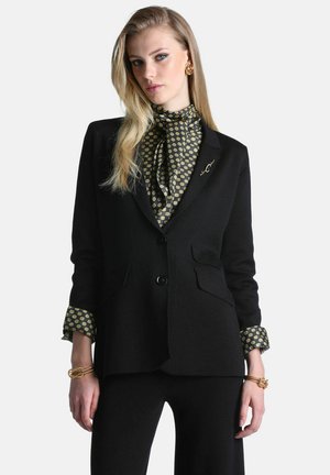 Femme aux longs cheveux blonds portant un costume noir, un chemisier à motifs, des boucles d'oreilles dorées et des bracelets sur un fond clair uni.