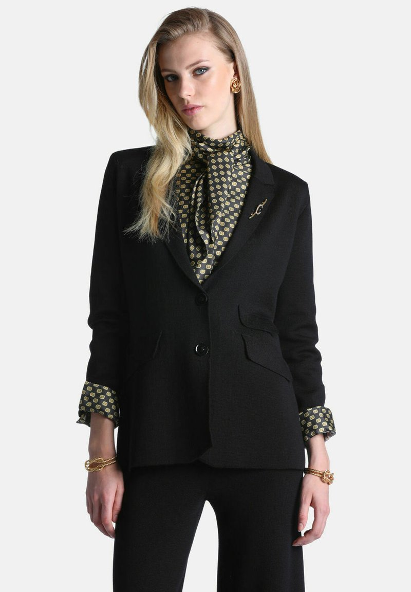 Femme aux longs cheveux blonds portant un costume noir, un chemisier à motifs, des boucles d'oreilles dorées et des bracelets sur un fond clair uni.