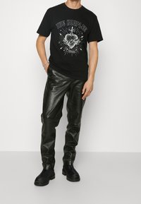 Camiseta de algodón negro con impresión gráfica que presenta un corazón y un dagger, combinada con pantalones de cuero negro sintético y botas negras hasta el tobillo.