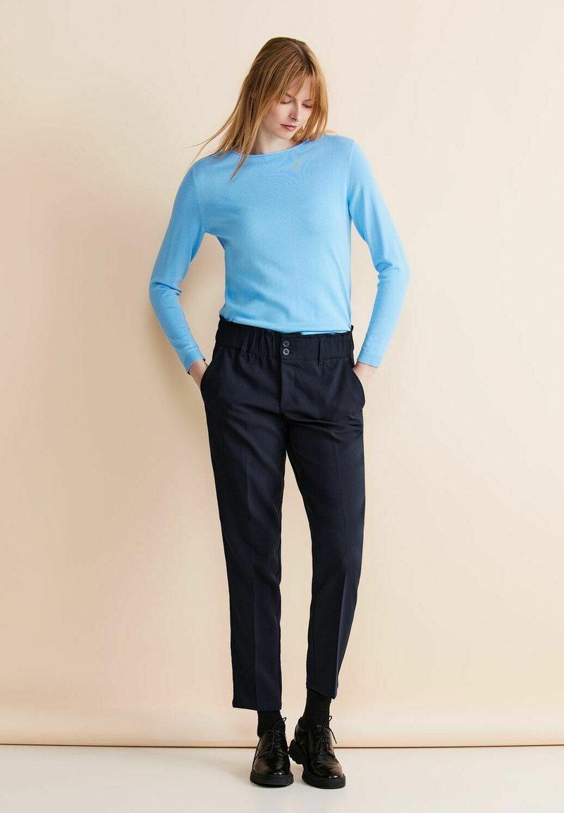 Ljusblå långärmad topp i kombination med mörkblå taperade byxor, med elastisk midja och framknappar, komplett med svarta skor.