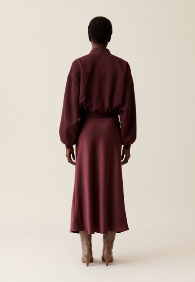 Haut bordeaux à manches longues avec manches ballons, associé à une jupe assortie. Texture lisse, longueur genoux, complété par des bottes à talons beiges clairs.