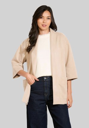 Frau mit langen, dunklen Haaren, die eine beige Jacke mit offenem Vorne, ein weißes Oberteil und dunkelblaue Jeans trägt, vor einem schlichten grauen Hintergrund steht.