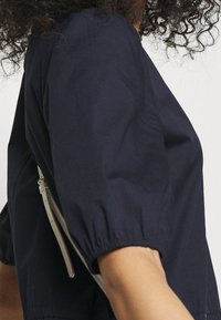 Mörkblå kortärmad blus med puffärmar, som har en mjuk textur och en strukturerad passform, kompletterad med en ljus färgad remaccessoar.