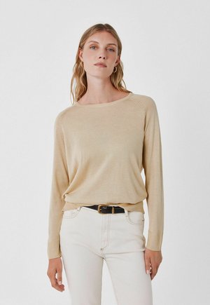 Vrouw met lang blond haar draagt een beige trui met lange mouwen, ingestopt in witte hoge taille broek met een zwarte riem, staand tegen een witte achtergrond.