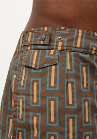 Bruine korte broek met geometrische patronen in teal en geel. Voorzien van een knoop en een klein achterzakje. Gemaakt van lichtgewicht stof.