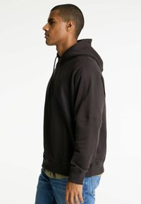 Hombre con sudadera con capucha negra y jeans azules de pie en perfil contra un fondo blanco.