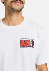 Camiseta de algodón blanca con un diseño gráfico rojo y negro en el lado izquierdo del pecho que dice "RVCA" y "EL EQUILIBRIO DE LOS OPUESTOS".