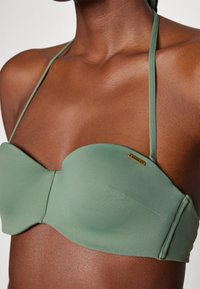 Grønn bandeau bikini topp med en glatt tekstur, tynne stropper og en liten gulllogo aksent på siden.