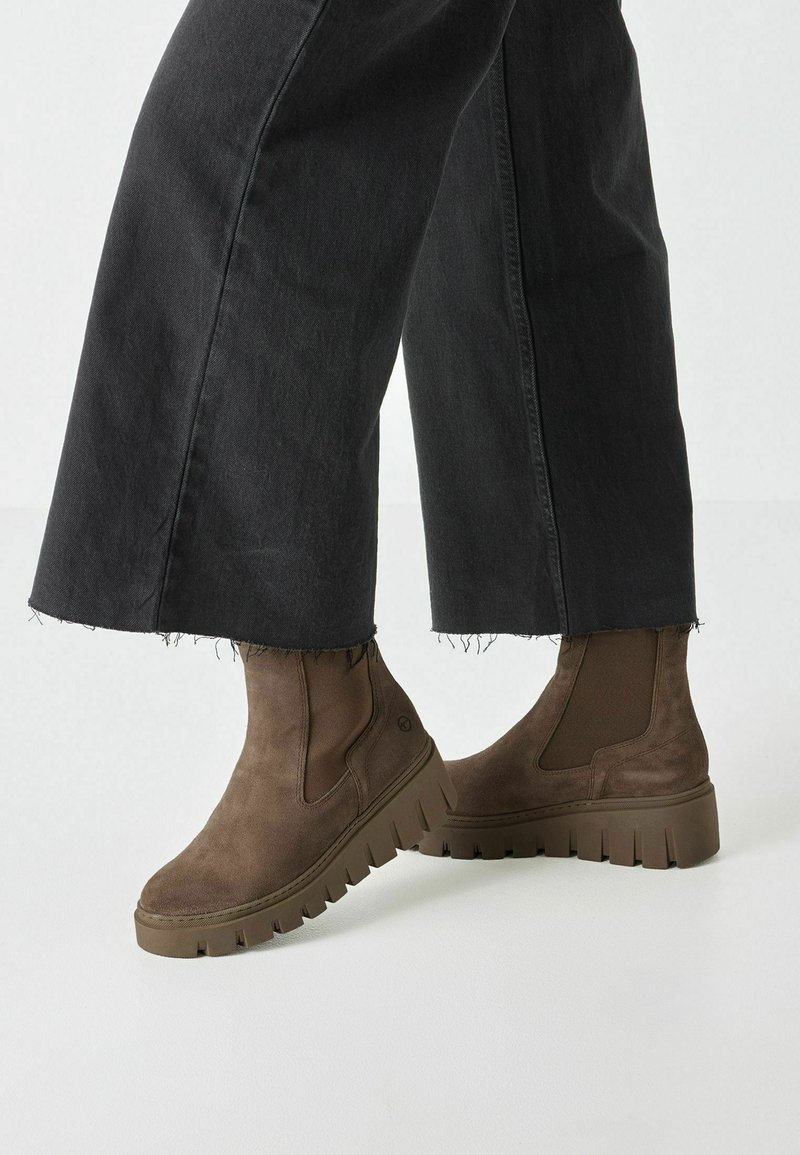Bottines en daim marron avec des panneaux latéraux élastiques, semelles texturées épaisses et un design épuré. Associées à un pantalon large noir.