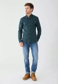 Camisa de color verde oscuro con botones dorados, combinada con jeans de color azul claro y botas de color beige. La camisa tiene una textura sutil y un bolsillo en el pecho.