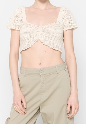 Kvinde iført en cremefarvet hæklet kortærmet croptop og højtaljede beige bukser, stående mod en ensfarvet baggrund.