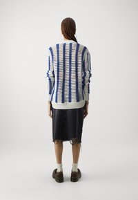 Cárdigan de punto con rayas azules y blancas, diseño holgado, puños acanalados, llevado sobre una falda de mezclilla oscura con dobladillo deshilachado. Zapatos marrones y calcetines blancos.